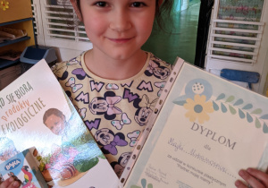 Zdjęcie przedstawia uśmiechniętą dziewczynkę która prezentuje dyplom i nagrodę za udział w konkursie plastycznym " Portret mojej mamy"